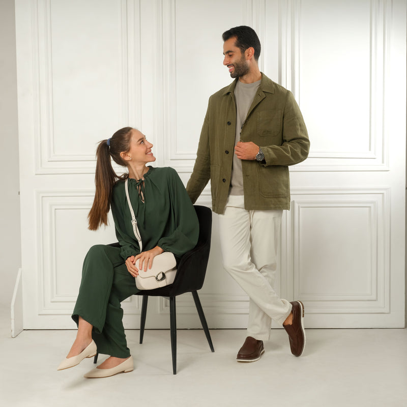 Woman in green outfit wearing cream flat pumps sits on a chair next to a man in casual attire against a white panel backdrop.