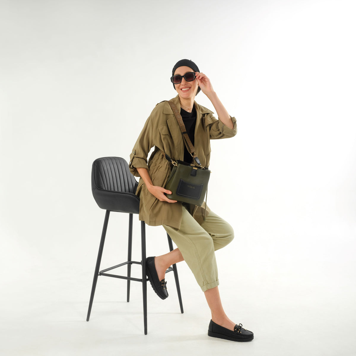 Woman sitting on stool wearing Adela moccasins flat in black, styled with a green outfit and sunglasses, showcasing a chic casual look.