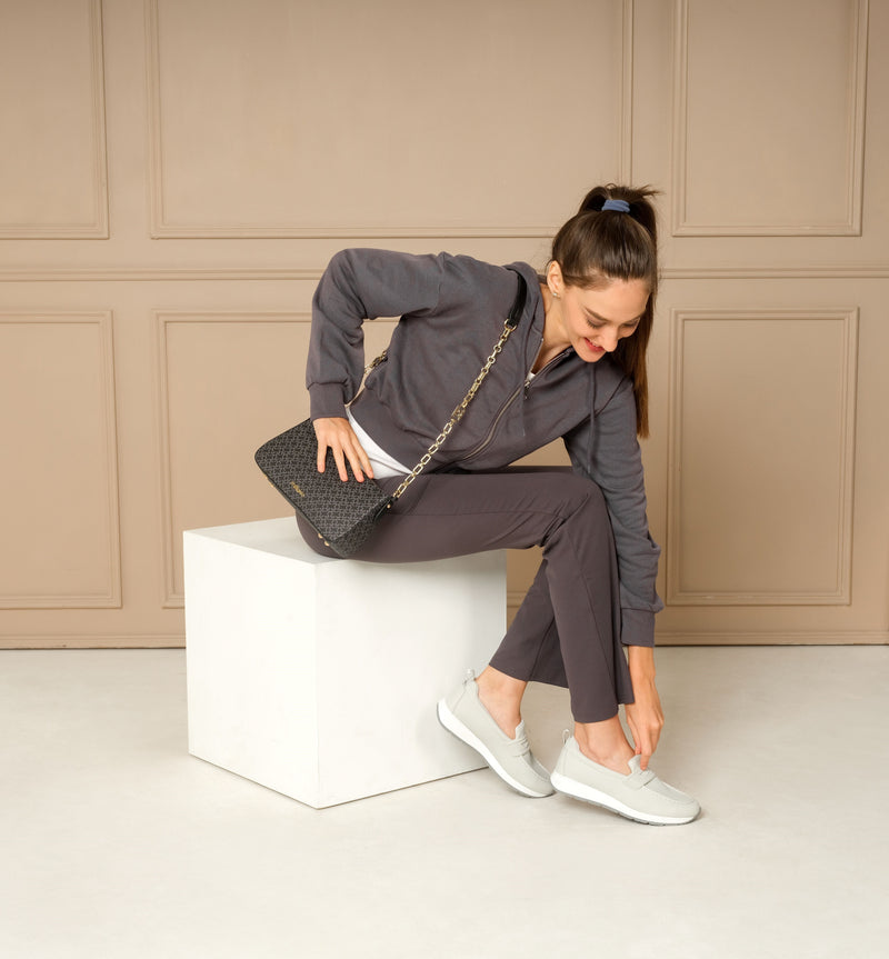 Woman wearing grey Marley moccasins with a sporty twist, featuring a strap detail and white midsole, sitting on a white cube.